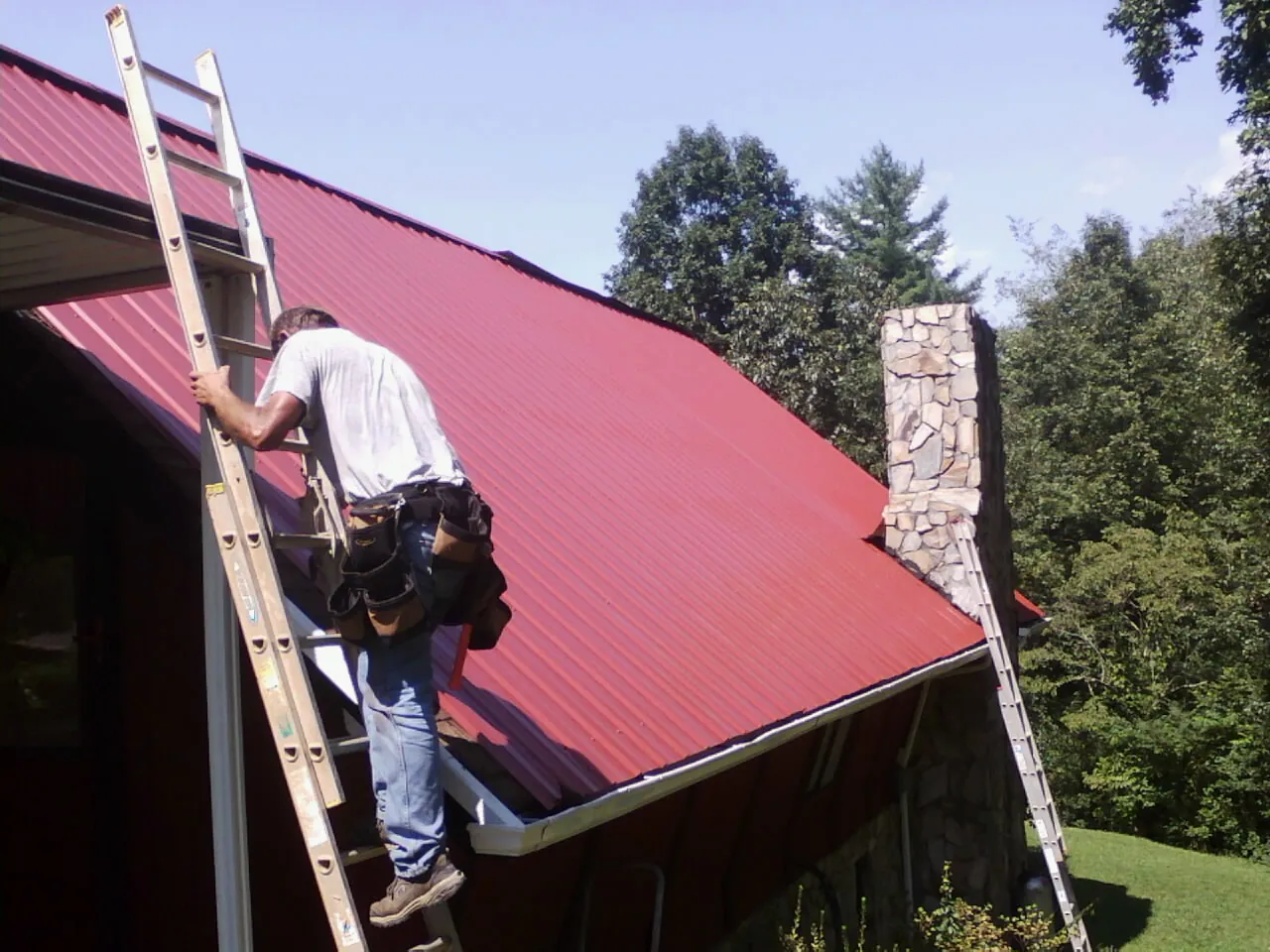 Roofer on a ladder beside a red metal roof and stone chimney — Soffit & Fascia Repair by Roofing Brothers in Beverly